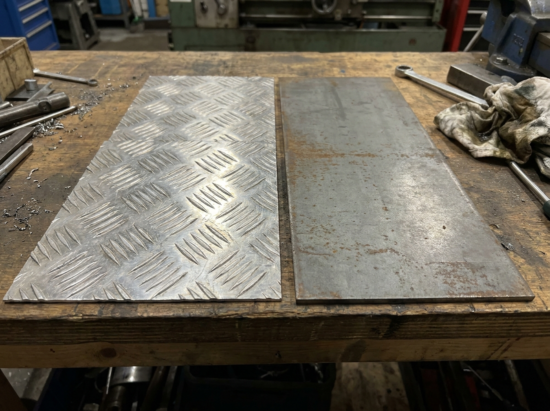 Aluminium chequer plate sheet beside a mild steel plate on a UK workshop bench — bright aluminium five-bar tread next to grey steel